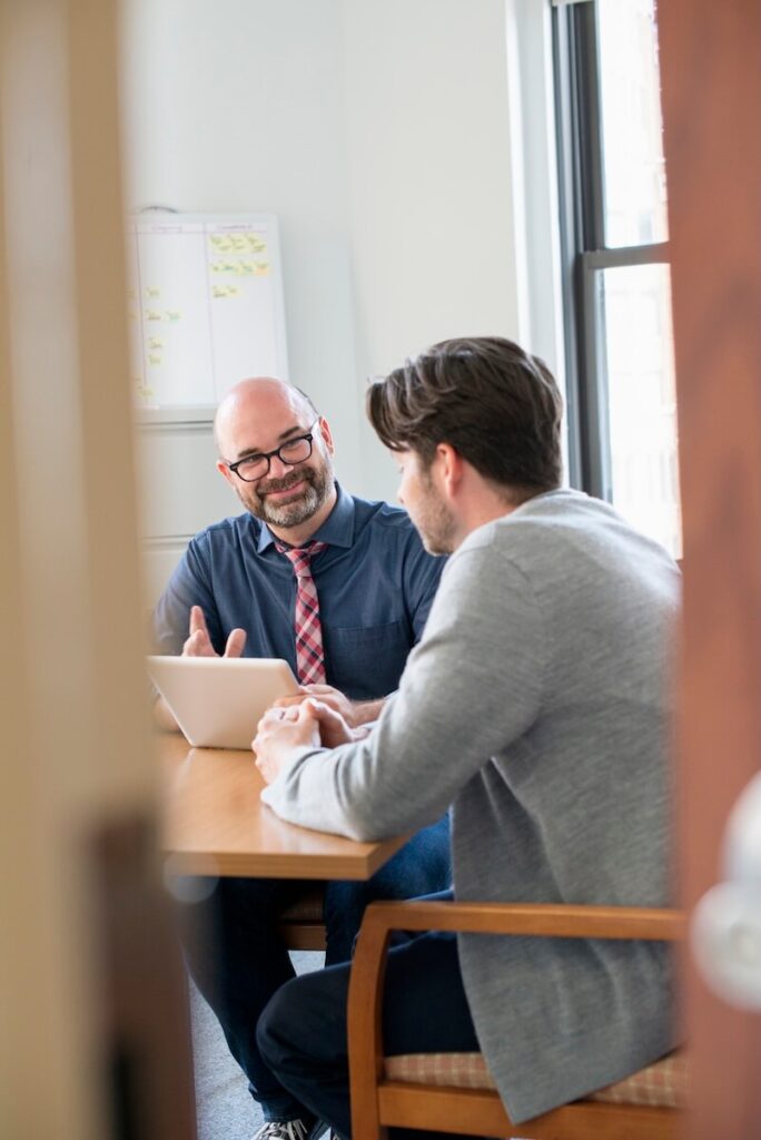 Two people are in an office, with one adult male talking while gesturing at a tablet. The man has glasses, a beard, and is wearing a blue collared shirt with a patterned tie. The other person is sitting and listening. There is a whiteboard on the wall.
